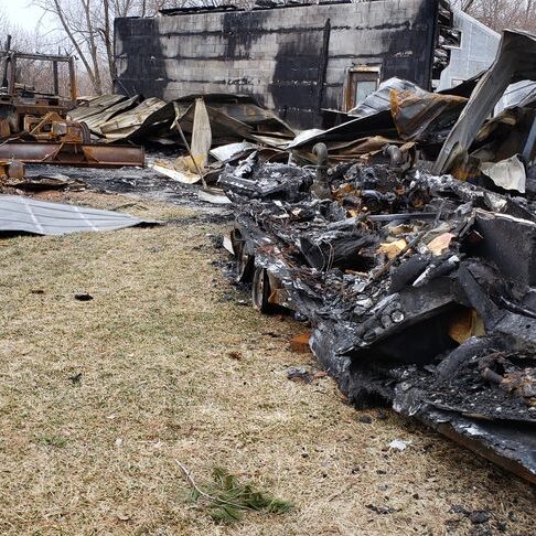 Charred debris and remnants of a burned structure scattered across a grassy area, illustrating the aftermath of a fire.